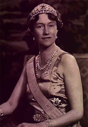 Grand Duchess Charlotte of Luxembourg wearing the Grand Duchess Adélaïde tiara. She's seated and smiling gently, wearing big diamond earrings, a diamond collet necklace, a silk sleeveless dress, the sash of an order across her chest, and diamond bracelets.