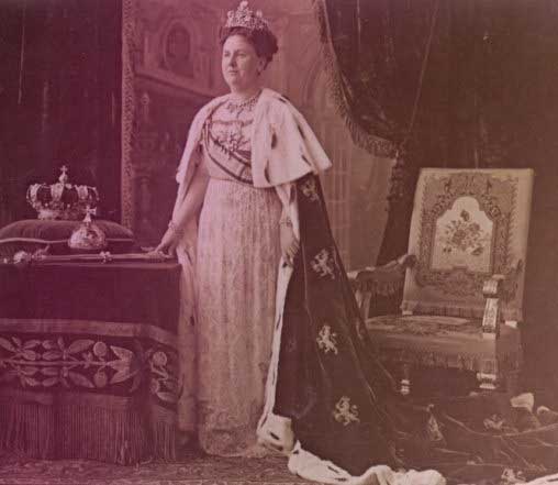 Queen Wilhelmina of the Netherlands in the Stuart tiara. She's standing in formal attire, with a long ermine-trimmed cape, with a crown, scepter, and orb on a table beside her.