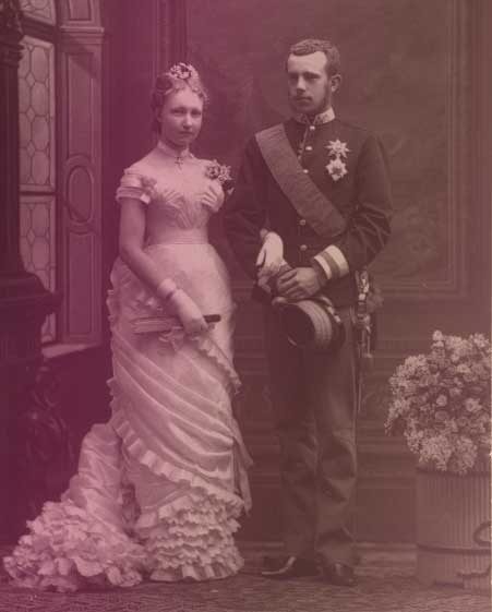 Stephanie with her new husband, Crown Prince Rudolf. They're standing arm-in-arm in formal dress. Staphanie wears an off-the-shoulder white ballgown, white gloves, and his holding a fan. Rudolf is wearing a military uniform and holding a hat in his hand.