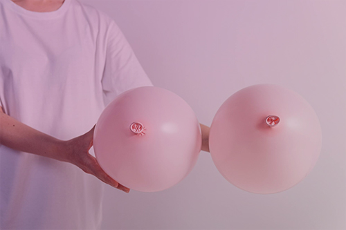 A person holding two pink balloons meant to represent breasts and symbolize the need for breast health exams.