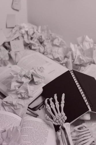 Books and crumpled paper cover a desk, with a skeleton’s hand visible in the bottom, stretched out over an overturned book.